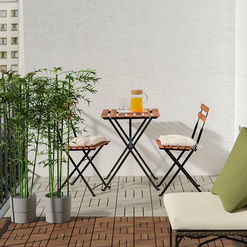 Acogedor balcón con terraza RUNDEN, pequeña mesa y sillas de madera, exuberante planta de bambú y fondo de pared blanca.