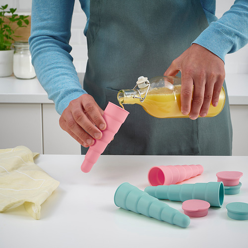 Una persona vierte jugo de naranja en coloridos moldes para paletas heladas en un mostrador de cocina, utilizando el producto UPPFYLLD.