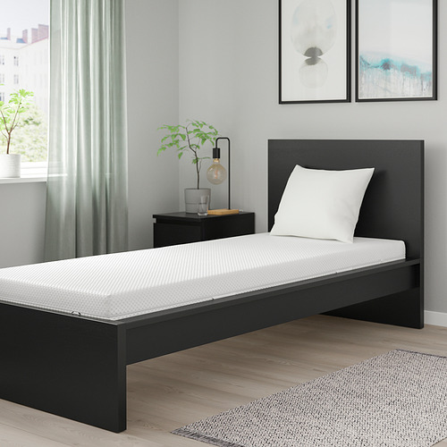 Dormitorio minimalista con cama individual ÅBYGDA negra, colchón blanco, almohada, suelo de madera clara y plantas.
