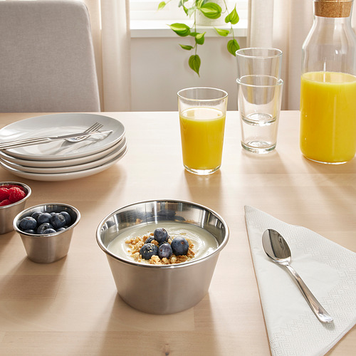 Juego de mesa con tazón de acero inoxidable con yogur, bayas y granola. Vasos de jugo de naranja y agua, platos, cubiertos y una planta.