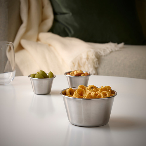 Tres tazones metálicos sobre una mesa blanca contienen bocadillos: pretzels, nueces mixtas y aceitunas verdes. Perfecto para reuniones al aire libre.