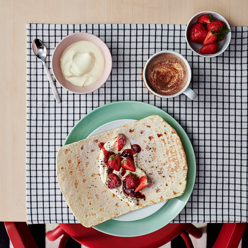 Una comida preparada sobre un mantel a cuadros con un crepe doblado con bayas, una taza de café, un tazón de crema y un tazón de fresas.
