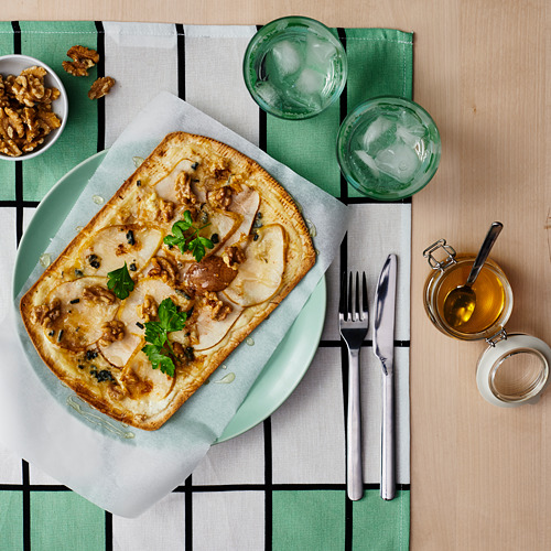 Un plato verde con pan plano cubierto con queso, nueces y hierbas acompañado de vasos de agua y un tazón de nueces sobre un mantel a cuadros verdes.