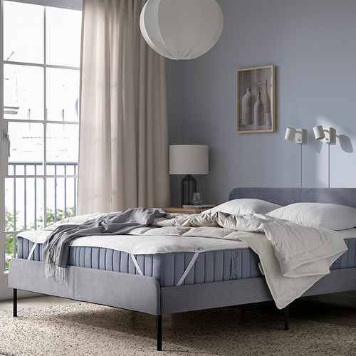 Dormitorio minimalista con cama gris sin hacer, ropa de cama blanca, lámpara de noche, cortina gris, pared azul claro y lámpara circular para colgar.