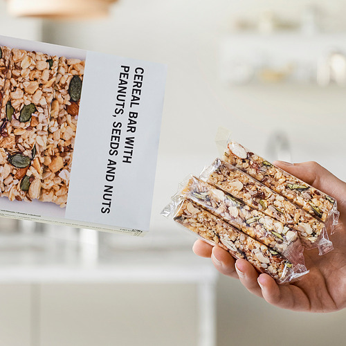 Sosteniendo con la mano tres barras de cereal envueltas junto a una caja que muestra una barra de cereal con imagen y texto con cacahuetes, semillas y nueces.