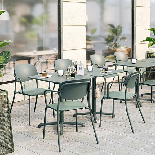 Moderna cafetería al aire libre con muebles VISINGSÖ . Mesas grises con ganchos, sillas apilables con reposabrazos y cojines, zona de azulejos, plantas, grandes ventanales.