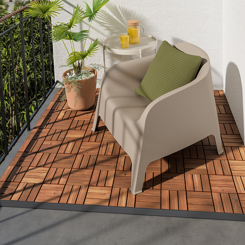 Acogedor balcón con silla RUNNEN beige, almohada verde, pequeña mesa redonda, dos bebidas amarillas y una gran planta en maceta en la terraza de madera.