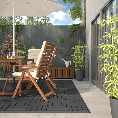 Patio con silla y mesa RUNNEN plegada en madera, cojines crema, sombrilla blanca, baldosas negras, casa gris, plantas exuberantes.