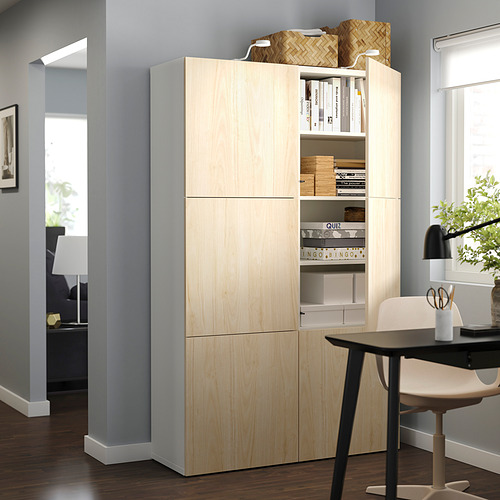 Un mueble de almacenaje BESTÅ de madera clara en una habitación. Tiene estantes ajustables y puertas parcialmente abiertas que revelan artículos organizados.