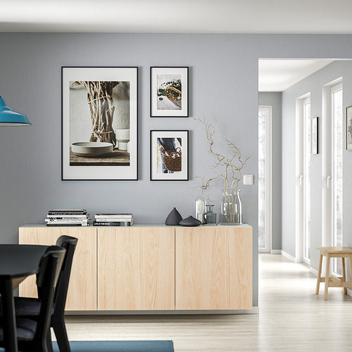 Sala de estar minimalista con gabinete BESTÅ de madera clara, detalles negros, jarrones, libros, cuadros enmarcados.