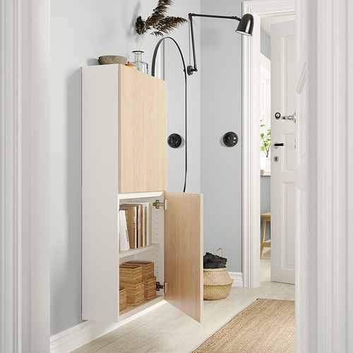 Pasillo moderno con almacenaje BESTÅ blanco. El armario abierto revela libros apilados y cesta de mimbre. Lámpara de pared negra y paredes gris claro.