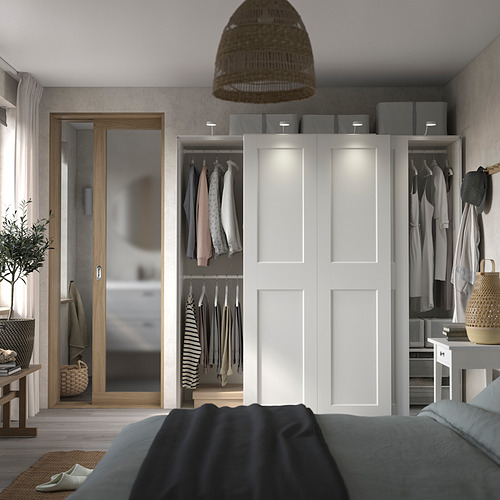 Dormitorio moderno con armario PAX blanco, ropa abierta, espejo, lámpara, planta.