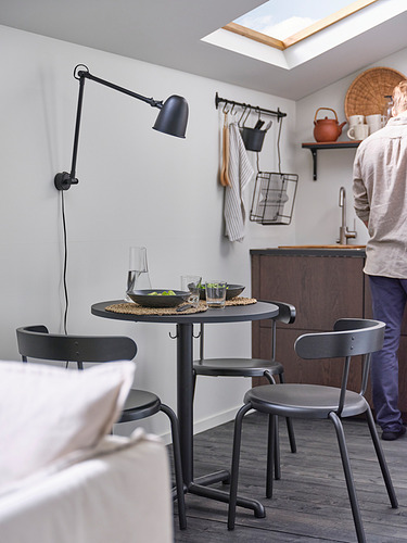 Rincón de cocina moderno con mesa redonda STENSELE negra, tres sillas, una lámpara de pared y una persona cocinando.