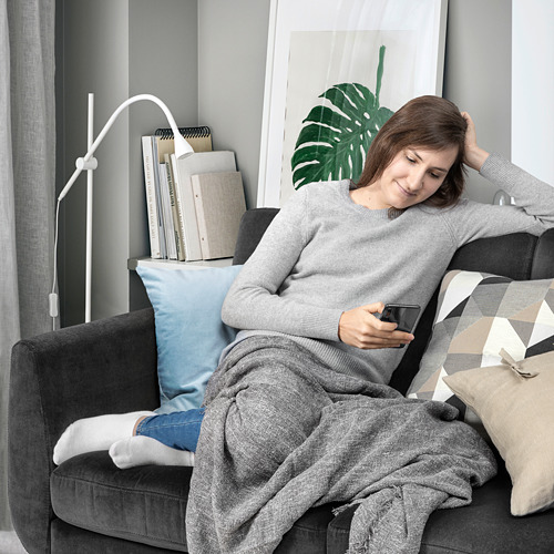 Una mujer se relaja en un sofá gris SMEDSTORP, usando su teléfono. Está abrigada bajo una manta, con una almohada azul y libros a su lado.