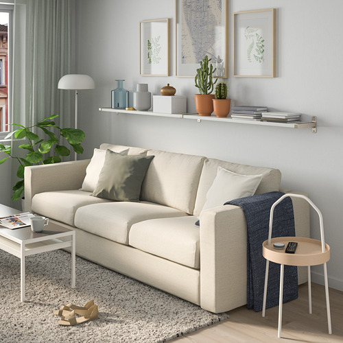 Sala de estar con estanterías BERGSHULT, jarrones, libros, plantas. Sofá crema con almohadas, manta azul, suelo de madera, mesas auxiliares.