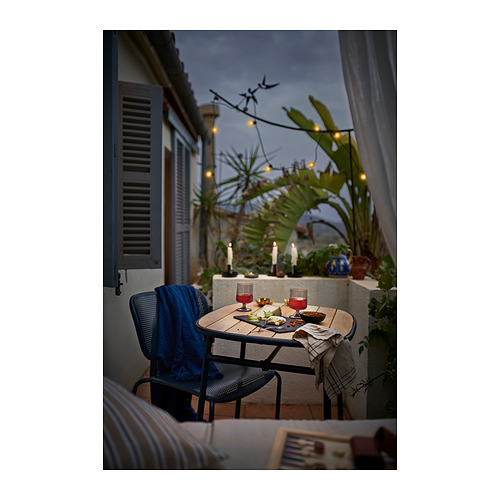 Mesa de balcón al aire libre con sillas DUVSKÄR negro-azul, cristalería y velas. Exuberante vegetación y guirnaldas de luces en el fondo.