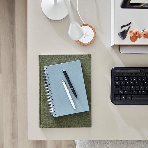 Un cuaderno azul y un bolígrafo sobre un escritorio luminoso con un teclado y una foto.