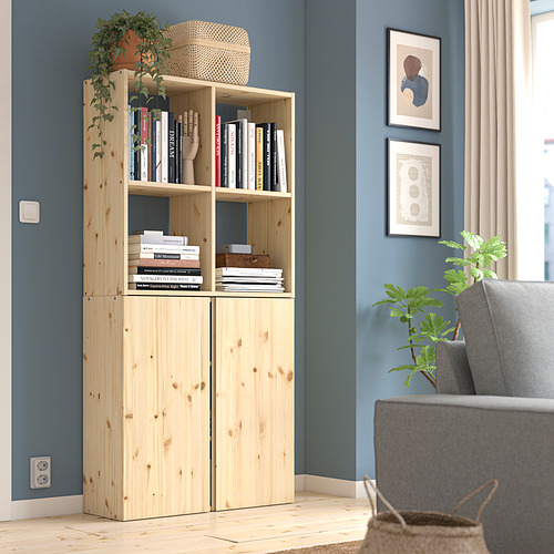 Librería IVAR de madera clara contra pared azul, llena de libros. Junto a una silla gris, una pequeña planta y cuadros enmarcados.