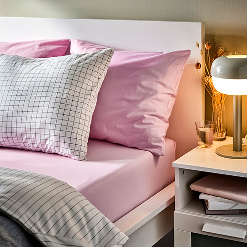 Cama ordenada con sábanas rosas, almohada de cuadros, lámpara, libros, plantas secas.
