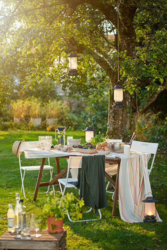 Montaje de fiesta en el jardín al aire libre debajo del árbol con mesa, sillas, linternas, cubiertos, platos, toallas y mantas.