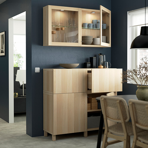 Mueble BESTÅ de madera clara con puertas y cajones de cristal contra la pared azul.