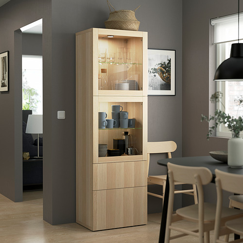 Mueble BESTÅ de madera clara con puertas de cristal, tazas y cuencos. Alto, delgado, encaja perfectamente en la esquina junto a las sillas de madera.