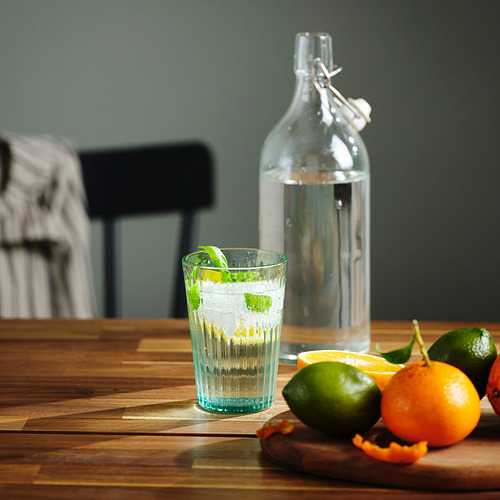 Un vaso lleno de agua y menta sobre una mesa de madera, con una botella y frutas cerca.
