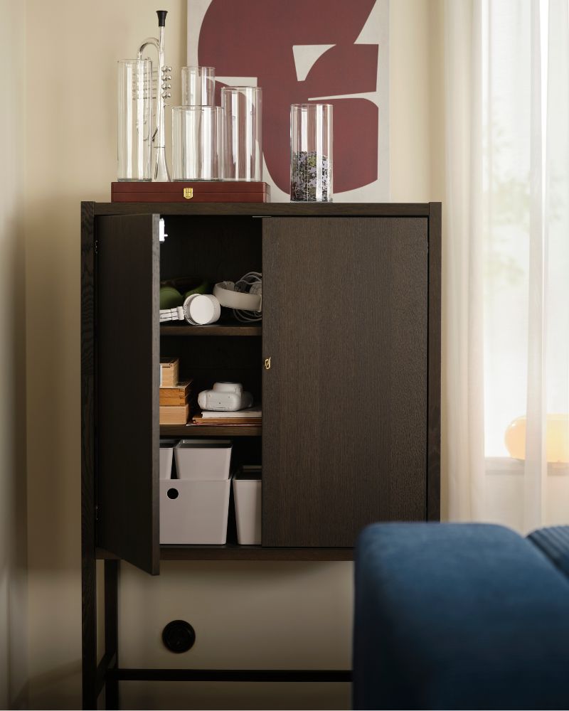 A living-room wall with a dark-brown STOCKHOLM 2025 cabinet holding various accessories by a window with sheer curtains.