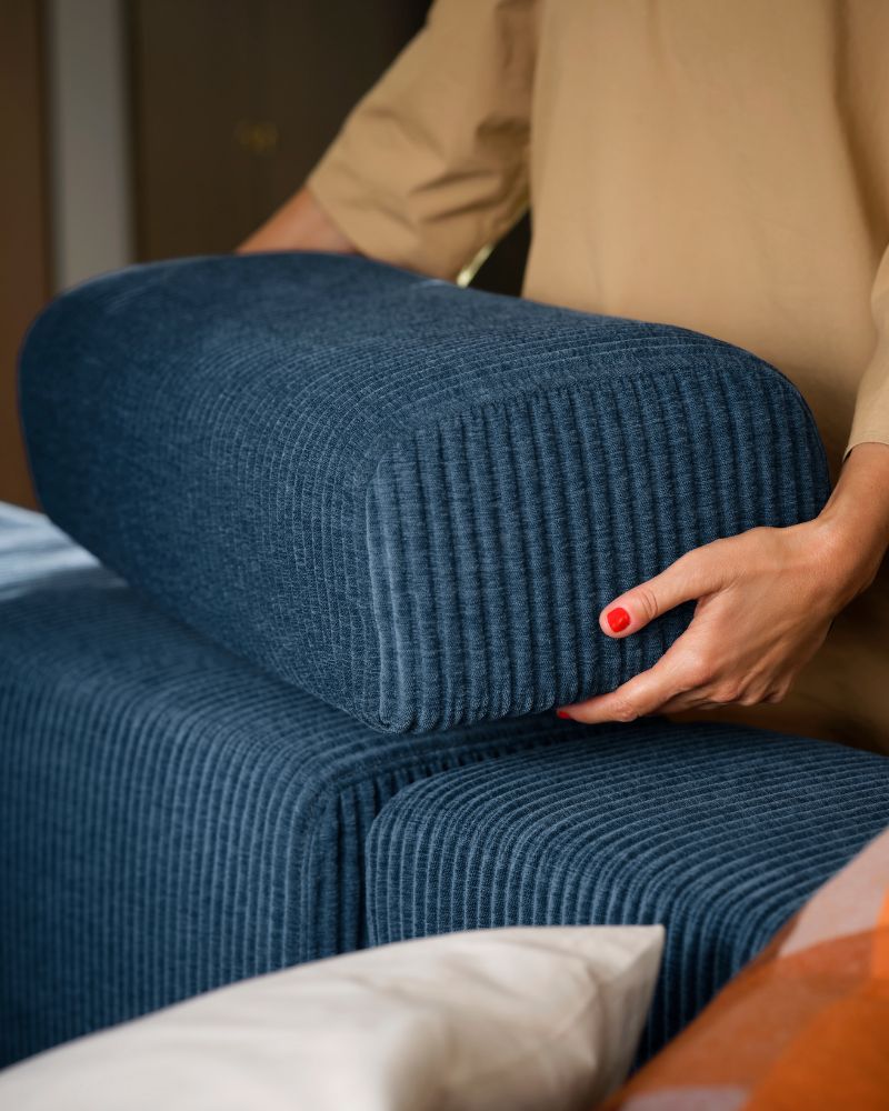 A person standing by a dark-blue JÄTTEBO four-seat modular sofa places a loose headrest on top of the backrest.