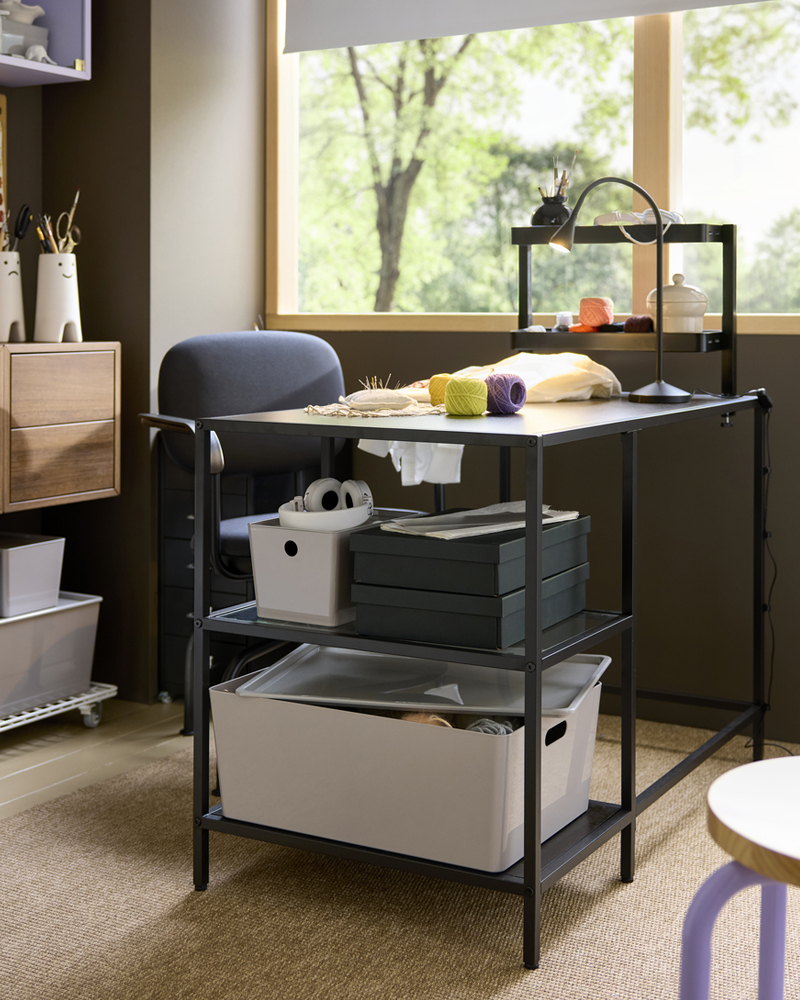 A workspace setup with a black-brown VITTSJÖ desk featuring a glass top, furnished with storage boxes and craft supplies.