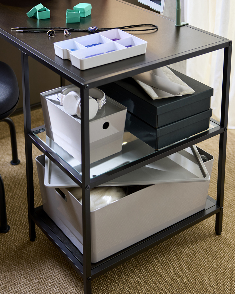 A close-up on a workspace setup with a black-brown VITTSJÖ desk with glass shelves, storing boxes and crafting supplies.