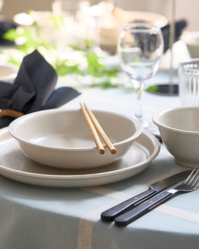 A dotted/light beige DAGGASTER bowl is placed on a plate, with two chopsticks on top of the bowl and cutlery on the side.