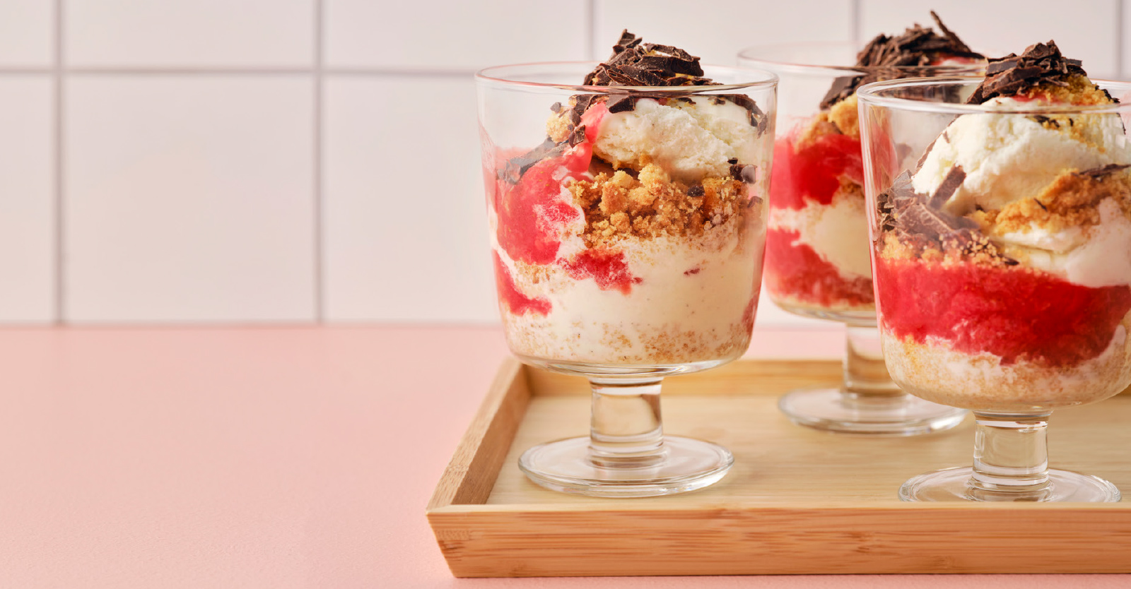 Three glass dessert cups filled with layered ice cream, red fruit coulis, crumbled biscuits, and topped with chocolate shavings on a wooden tray.