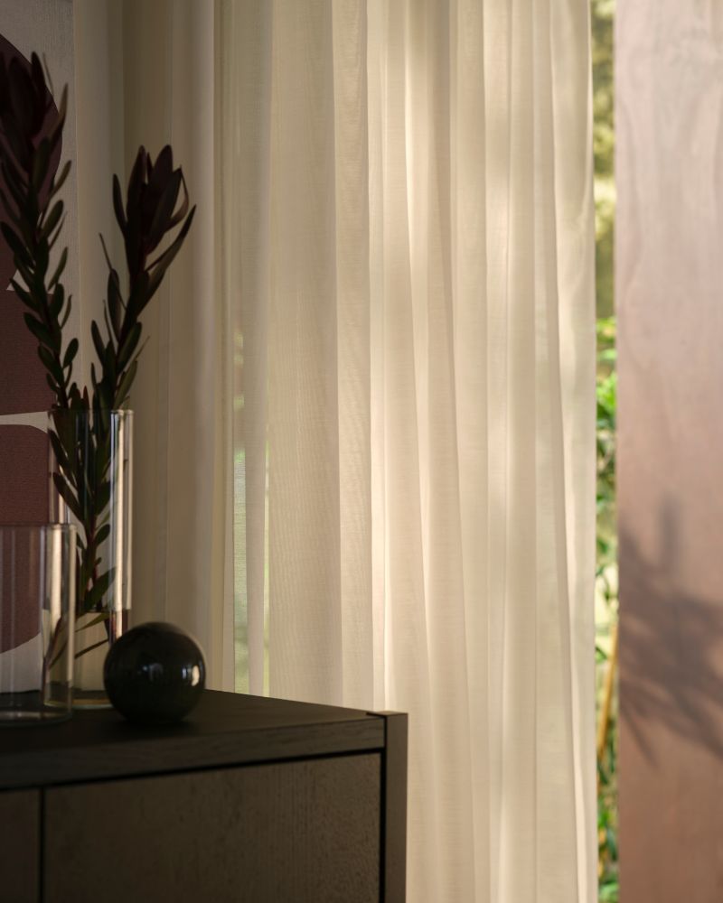 A living-room corner with a window covered by white STENFRÖ sheer curtains next to a dark-brown STOCKHOLM 2025 cabinet