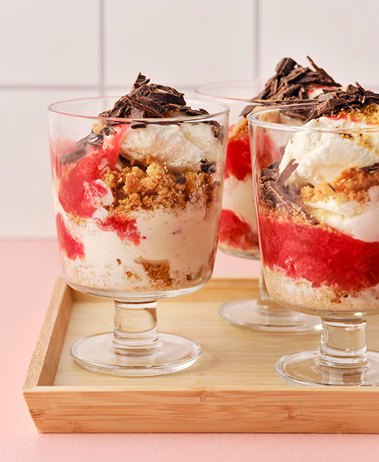 Three glass dessert cups filled with layered ice cream, red fruit coulis, crumbled biscuits, and topped with chocolate shavings on a wooden tray.