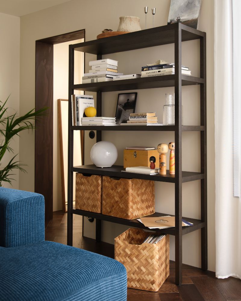 A living room with a dark-brown oak-veneer STOCKHOLM 2025 shelving unit. It is filled with decor, books, and baskets.