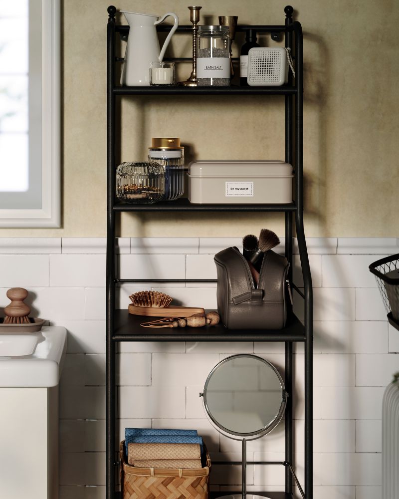 A black metal FRÖSJÖN shelving unit holds toiletries, decorative items and hand towels in a traditional-styled bathroom.