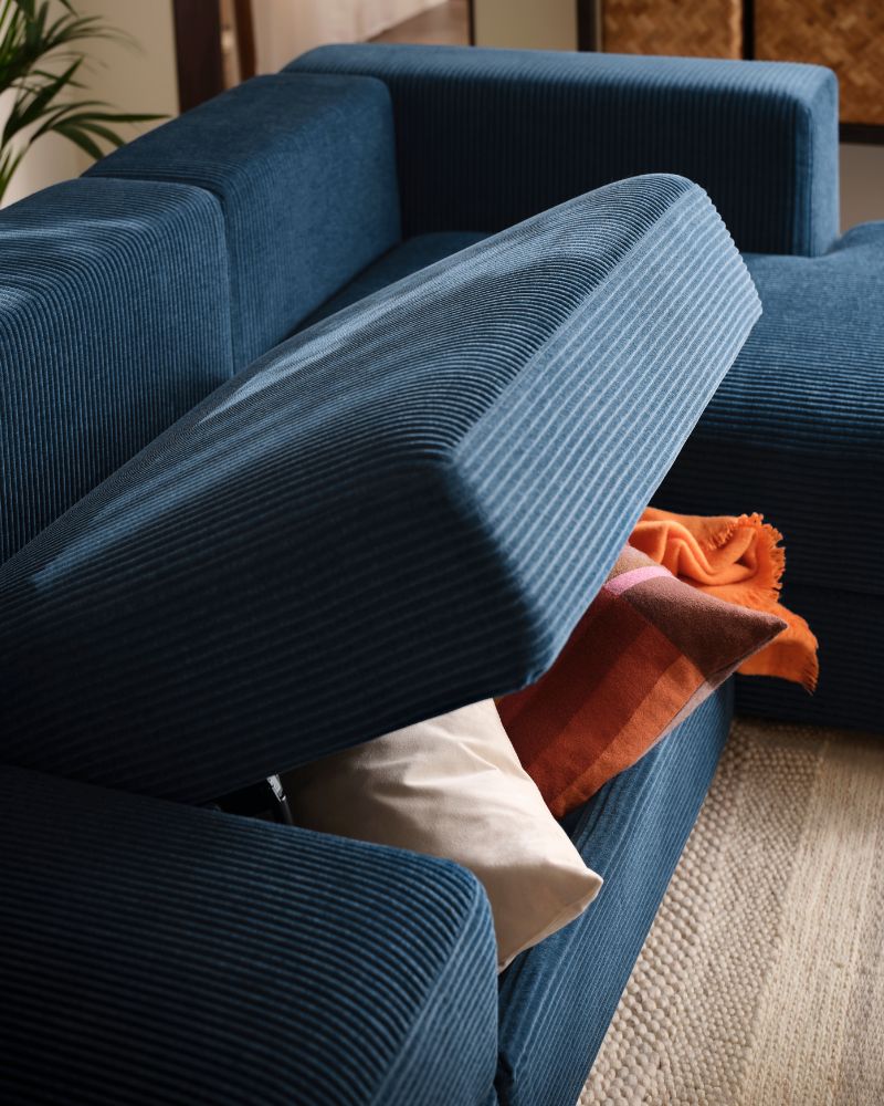 A living room with a dark-blue JÄTTEBO four-seat sofa, the middle seat raised, holding a pair of cushions and a throw within.