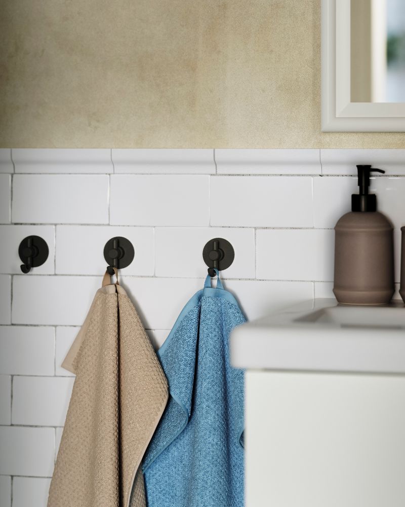 A dark blue-grey GULVIAL hand towel hangs on one of three hooks on a tiled wall next to a white wash-basin in a bathroom.
