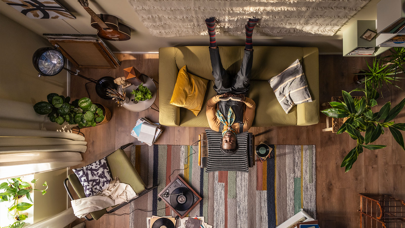 Overhead view: person with bunny ears, upside down on green sofa, cozy living room with plants, guitar, and record player.