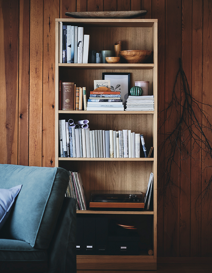 Estantería de madera con libros, cuencos decorativos y un tocadiscos, sobre fondo de pared de madera, junto a un sofá azul.
