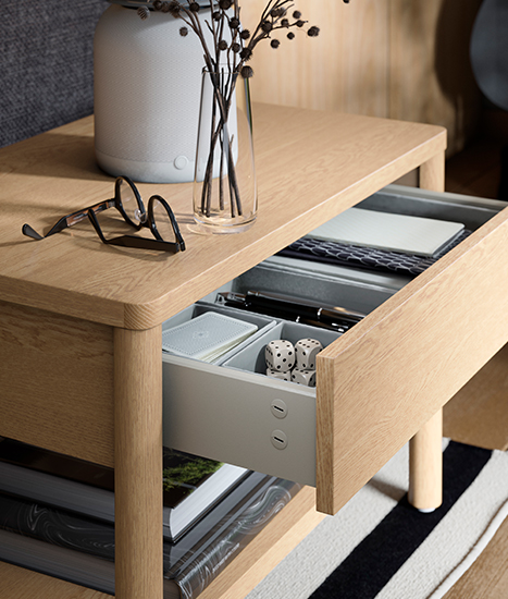 Mesa auxiliar de madera clara con cajón abierto que muestra organizadores con pequeños objetos, junto a un jarrón decorativo y unas gafas.