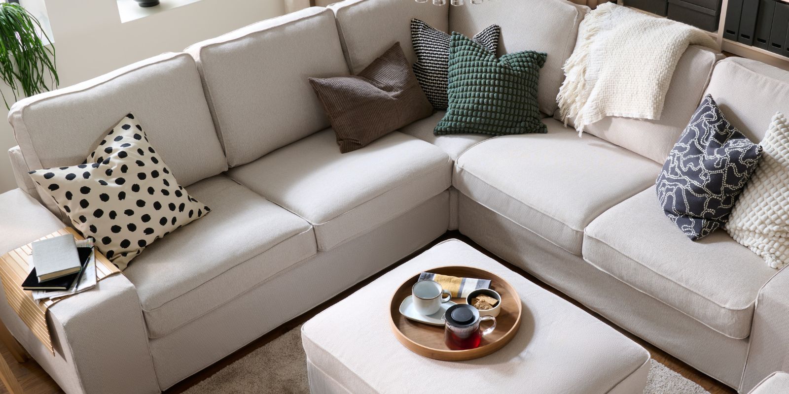 A light, bright living room with a KIVIK corner  sofa and with colourful cushions, a throw and a  centrally placed footstool.