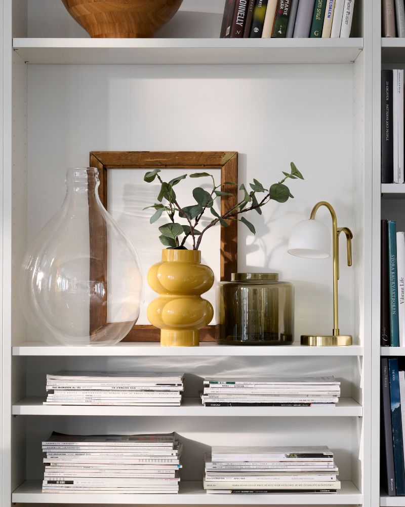 Neatly organised white BILLY bookcases  with a yellow-beige PÄRLHÄGG vase and a  TVÅMASTAD portable lamp sharing a shelf.