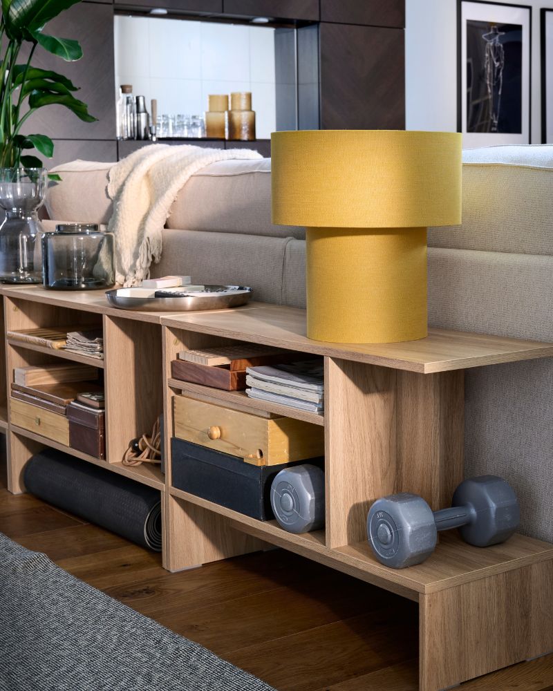 HOLMERUD sidetables with mixed contents  and a yellow STOCKHOLM table lamp atop are  placed along the back of a KIVIK sofa.