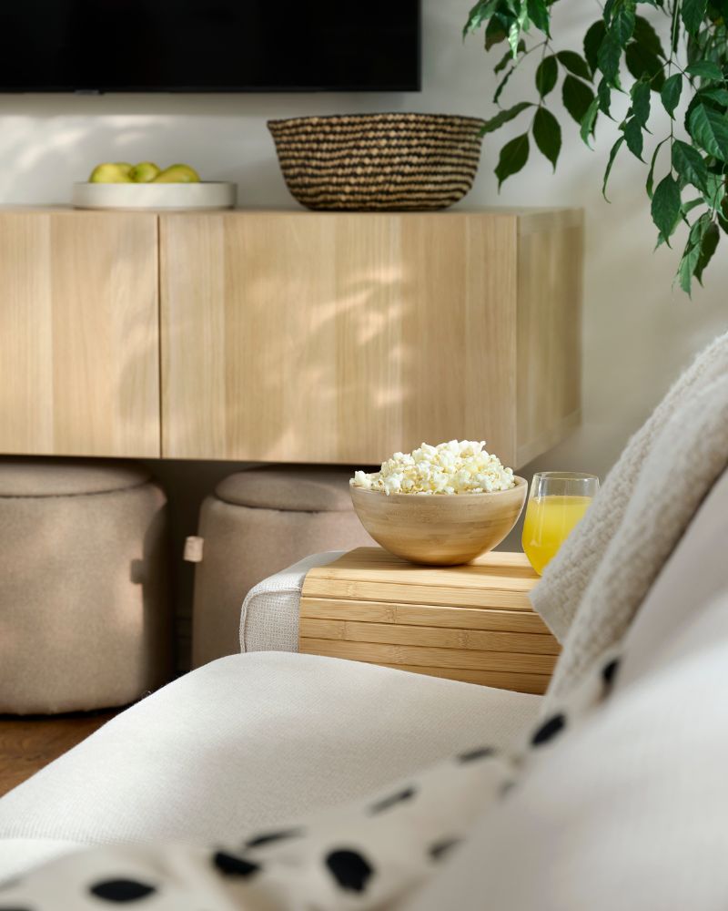Snacks and a glass of juice rest on a RÖDEBY  armrest tray placed the end of a KIVIK sofa in  a bright living room.