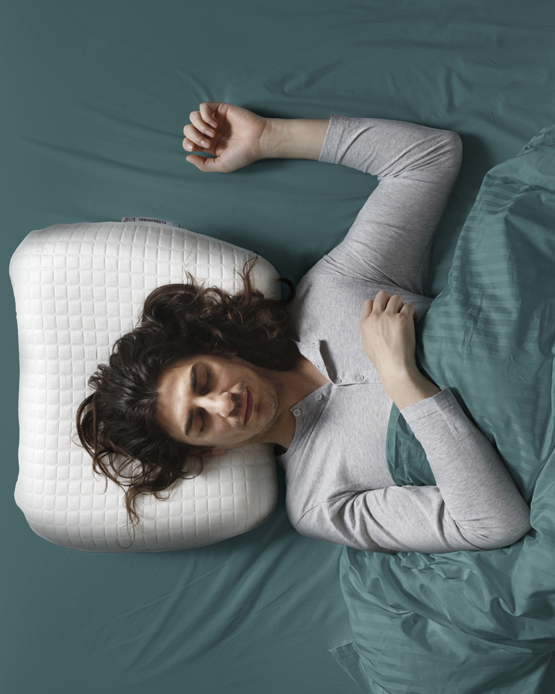 A bird’s eye view of a person asleep on their back with their arm up and their head on a KLUBBSPORRE ergonomic pillow.