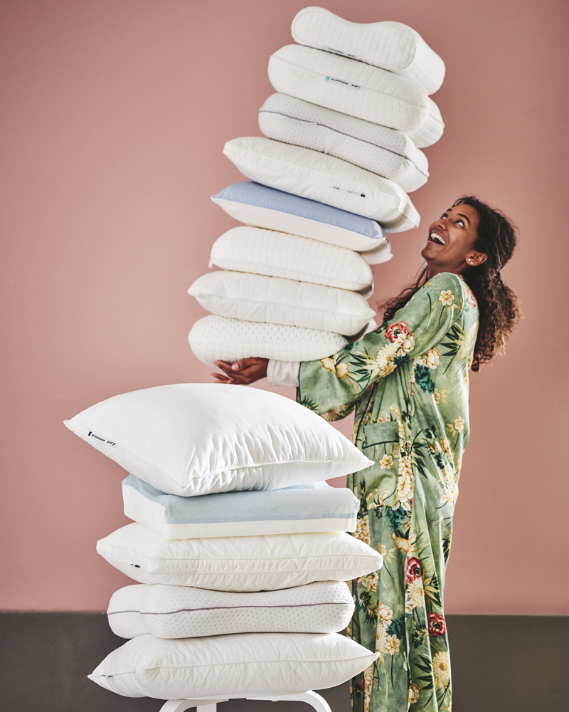 A person holds a high pile of various ergonomic pillows. A smaller pile is in front with a SKOGSFRÄKEN pillow on top.