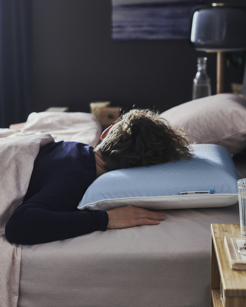 In a dark bedroom, a person in blue pyjamas in bed with a KVARNVEN ergonomic pillow is sleeping on their stomach.