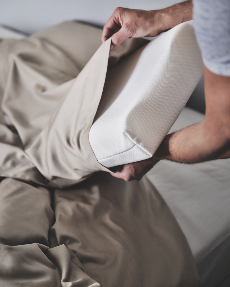Two hands holding white PAPEGOJBUSKE ergonomic pillow for side/back sleeper and calming beige satin bedlinen.